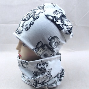 Könnte beinhalten: Ein weißes Mützen- und Halstuch-Set mit einem schwarz-grauen Roboter-Design. Die Mütze hat einen umgeschlagenen Rand. Das Halstuch ist röhrenförmig. Das Set ist aus einem weichen Stoff gefertigt.