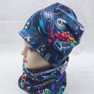 Könnte beinhalten: Dunkelblaues Mütze- und Halstuch-Set mit einem lebendigen Aufdruck von Spielcontrollern, Computermäusen und anderen Gaming-Grafiken. Die Mütze hat einen umgeschlagenen Rand, und das Halstuch ist röhrenförmig. Das Set wird auf einem Schaufensterkopf präsentiert.