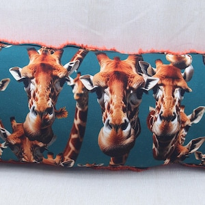 Könnte beinhalten: Rechteckiges Zierkissen mit türkisblauem Hintergrund und einem sich wiederholenden Muster von Giraffengesichtern. Die Giraffen haben braune und orangefarbene Markierungen. Das Kissen hat eine korallenfarbene Fransenborte.