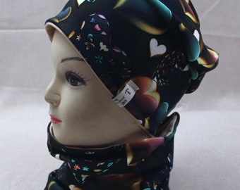 May include: A black beanie and neck warmer set with a colorful heart pattern. The beanie has a beige lining and the neck warmer has a black lining. Both items have a small tag with the word "Lie" on them.