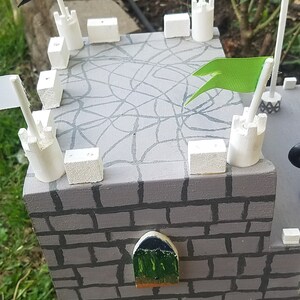 May include: A gray wooden castle with white towers and green flags. The castle has a brick wall and a small window. The roof is decorated with a spiderweb design.