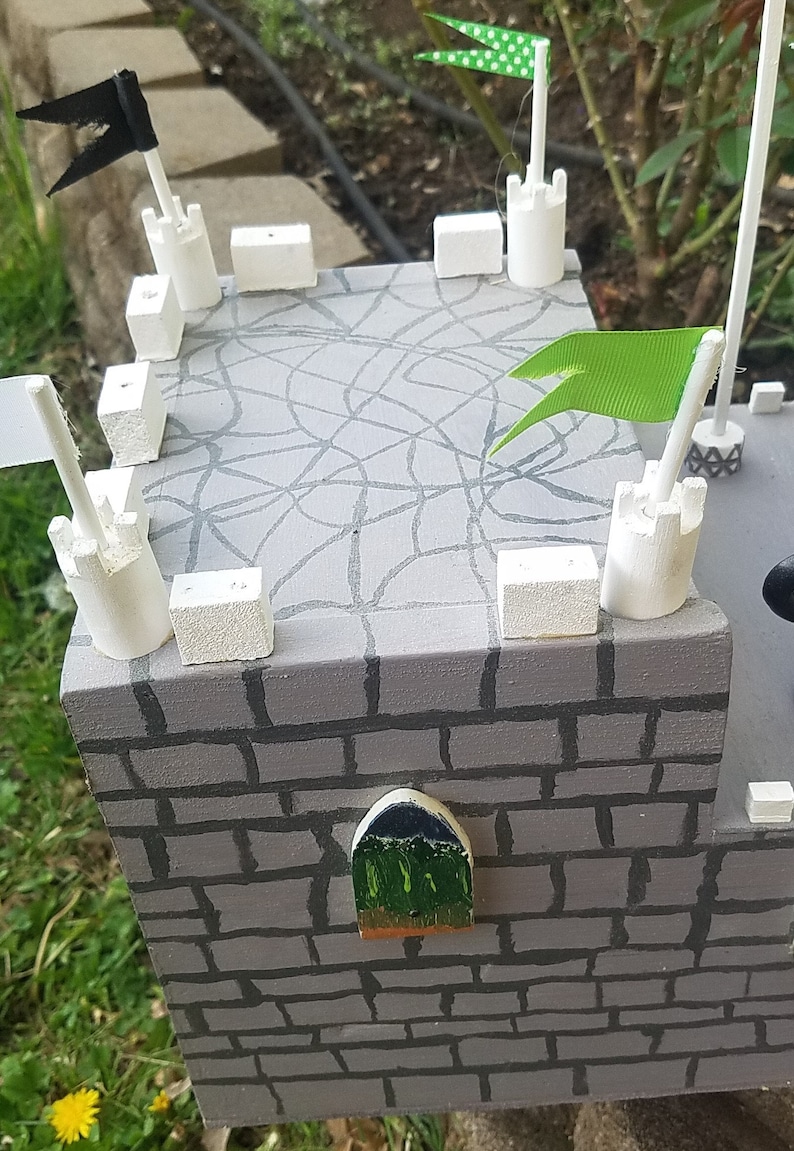 May include: A gray wooden castle with white towers and green flags. The castle has a brick wall and a small window. The roof is decorated with a spiderweb design.