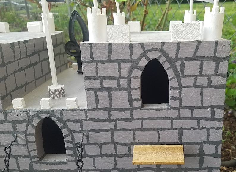 May include: A gray wooden castle model with white painted details, including towers and a small wooden bridge. The castle has two arched windows and a gray brick pattern.