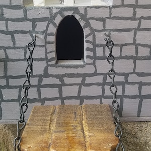 May include: A wooden drawbridge with black metal chains hangs from a gray stone castle wall. The drawbridge is raised and the chains are hanging loose.