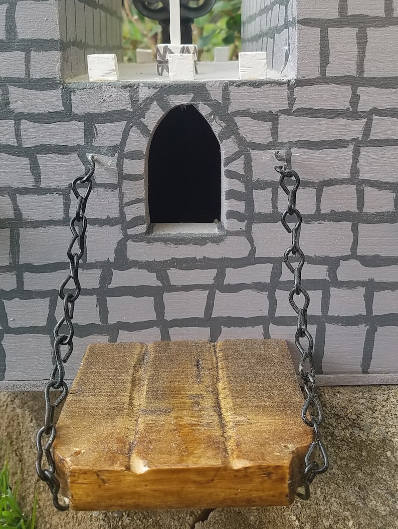 May include: A wooden drawbridge with black metal chains hangs from a gray stone castle wall. The drawbridge is raised and the chains are hanging loose.