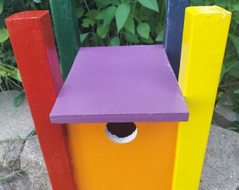May include: A brightly painted birdhouse with a vibrant color scheme. The birdhouse features a purple roof, an orange body, and is accented with red, green, yellow, and blue supports. The birdhouse has a circular entrance hole.