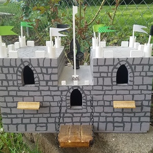 May include: A gray wooden castle toy with white towers and black and green flags. The castle has a drawbridge and windows.