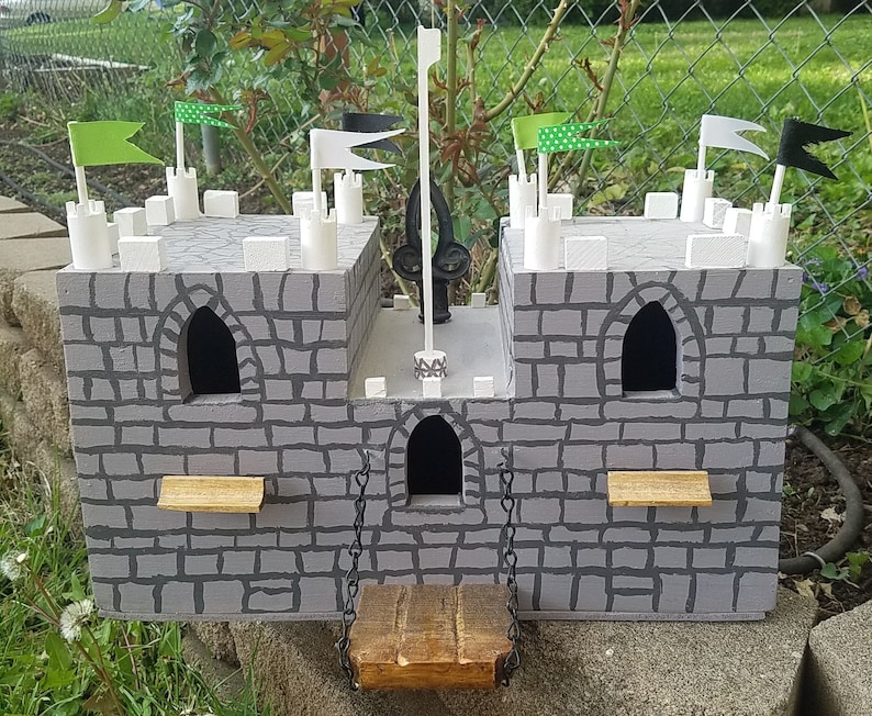 May include: A gray wooden castle toy with white towers and black and green flags. The castle has a drawbridge and windows.