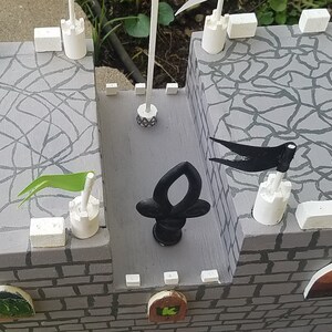 May include: A gray wooden castle playset with white towers and black flags. The castle has a gray stone wall with a brick pattern and green grass.