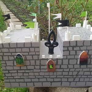 May include: A gray wooden castle toy with white towers and black and green flags. The castle has three small doors with green, brown, and a green and black design. The castle has a black metal finial on top.