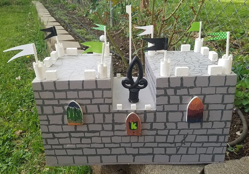 May include: A gray wooden castle toy with white towers and black and green flags. The castle has three small doors with green, brown, and a green and black design. The castle has a black metal finial on top.