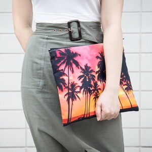 May include: A woman is holding a clutch purse with a palm tree print against a white tiled background. The purse has a pink and orange sunset background with black palm trees.