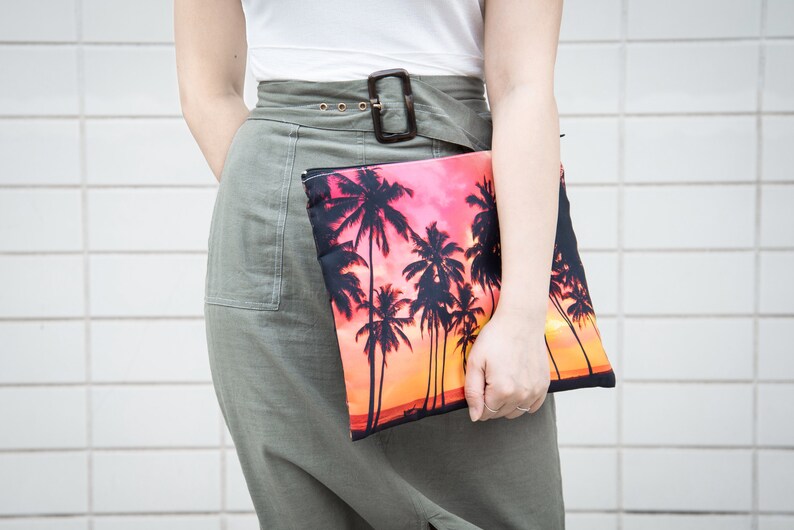 May include: A woman is holding a clutch purse with a palm tree print against a white tiled background. The purse has a pink and orange sunset background with black palm trees.