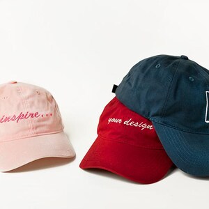 May include: Three baseball caps, one pink with the text "inspire..." embroidered in pink, one red with the text "your design" embroidered in white, and one dark blue with a white letter "K" embroidered on the side.