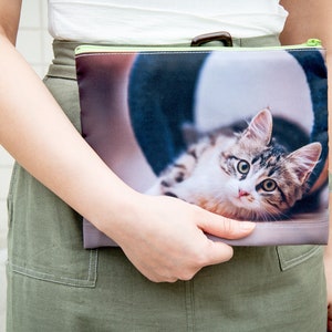 May include: A green zippered pouch with a photo of a tabby cat printed on it. The cat is lying down and looking at the camera.
