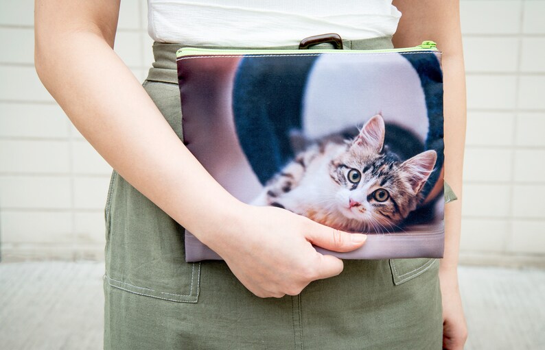 May include: A green zippered pouch with a photo of a tabby cat printed on it. The cat is lying down and looking at the camera.