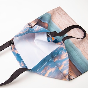 Peut inclure: Un sac fourre-tout avec des sangles noires et une scène de plage de coucher de soleil colorée imprimée sur le tissu. Le sac est ouvert et l'intérieur est blanc.