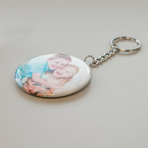 Pode incluir: Um chaveiro redondo com uma foto de duas crianças se abraçando. O chaveiro é prateado e tem uma corrente prateada.