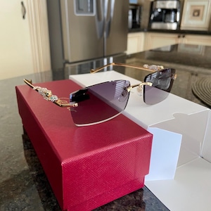 Puede incluir: Gafas de sol sin montura con montura dorada y lentes degradadas. Las patillas están adornadas con adornos decorativos. Las gafas de sol descansan sobre una caja roja texturizada y una caja blanca.