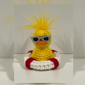May include: A handmade yellow crochet duck wearing silver glitter sunglasses and a red and white life preserver. The duck has a yellow tuft of hair on top of its head. The duck is a whimsical, decorative item.