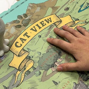 May include: A detailed map with a yellow banner reading "CAT VIEW" is shown. The map features green and tan terrain with illustrations of trees, mountains, and buildings. A hand is resting on the map, partially obscuring the details. The map has a whimsical, illustrated style.