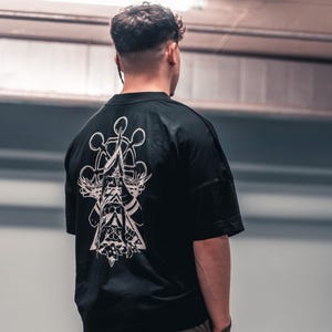 May include: A black t-shirt with a white graphic design on the back. The design features a stylised image of a hand holding a compass with a triangle and other geometric shapes.