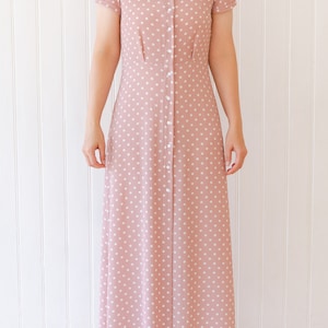 Może przedstawiać: Długa, różowa sukienka w białe kropki z zapięciem na guziki z przodu. Sukienka ma krótkie rękawy i kołnierzyk.