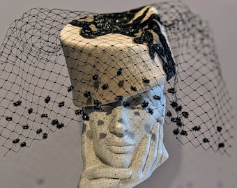 May include: A beige hat with black trim and a black mesh veil. The hat has a wide brim and a rounded crown. The veil is attached to the hat with a black ribbon.