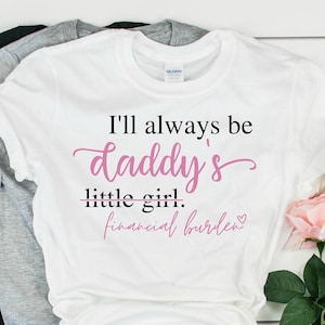 Puede incluir: Camiseta blanca con el texto "I'll always be daddy's little girl. financial burden" en letras negras y rosas. La camiseta está doblada y se muestra sobre una superficie de madera blanca con un par de zapatillas blancas y un ramo de rosas rosas y blancas.