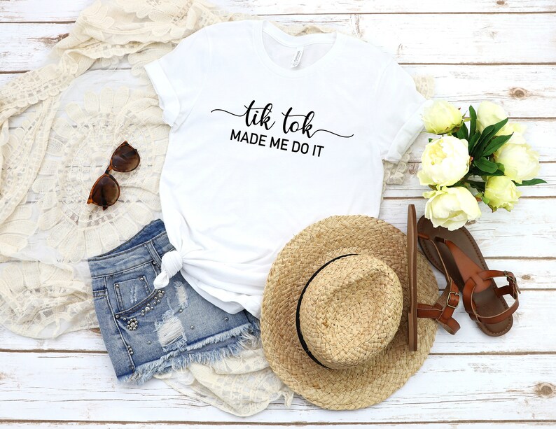 May include: White t-shirt with the text "tik tok made me do it" in black cursive lettering. The shirt is laid out on a white wooden surface with a straw hat, denim shorts, sunglasses, and white flowers.