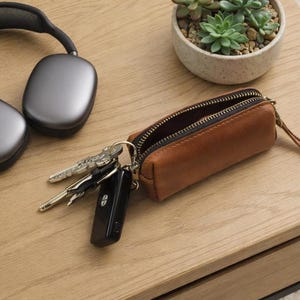 Könnte beinhalten: Ein braunes Leder-Schlüsseletui mit Reißverschluss, das mehrere Schlüssel enthält. Das Etui ist geöffnet und gibt den Blick ins Innere frei. Daneben befinden sich silberne Kopfhörer und eine kleine Sukkulentenpflanze in einem Topf auf einer Holzoberfläche.