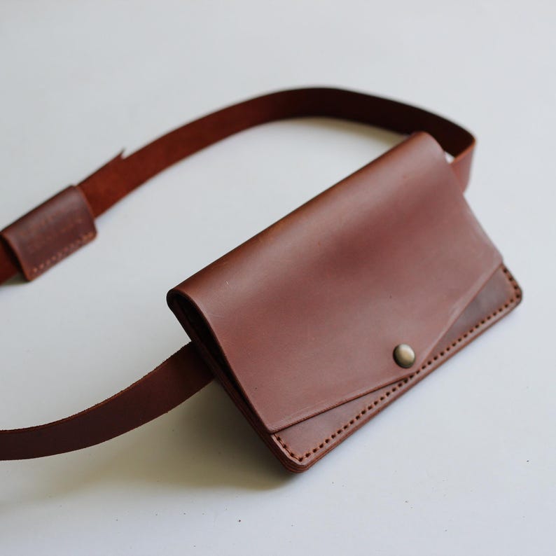 May include: A brown leather belt bag with an adjustable strap. The bag has a flap closure secured by a metal popper. The leather has a smooth texture and is stitched along the edges.