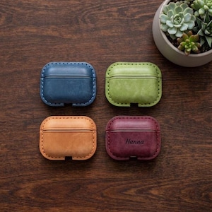May include: Four rectangular leather cases for wireless earbuds, each featuring visible stitching along the edges. The cases are coloured blue, light green, tan, and dark red. The dark red case has the name Hanna embossed on the front surface.