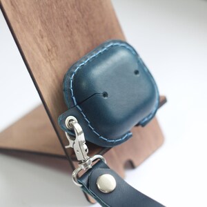 May include: A dark blue leather case with a light blue stitching detail for AirPods with a silver metal carabiner and a dark blue leather strap.