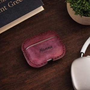 May include: A burgundy leather AirPod case with the name "Hanna" engraved on the front. The case has a stitched border and a silver metal accent. A pair of silver headphones is visible in the background.