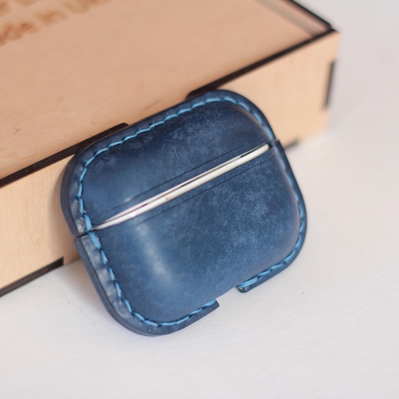 May include: A blue leather case with a silver metal trim for protecting AirPods. The case has a stitched edge and a distressed look.