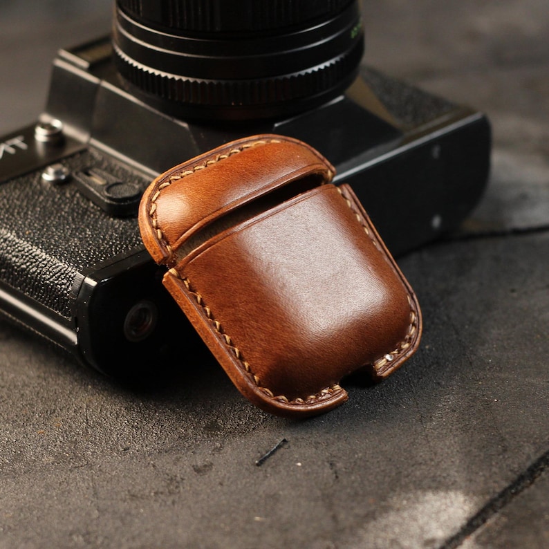 May include: A brown leather case for AirPods, with a stitched seam around the edge. The case is sitting on top of a vintage camera.