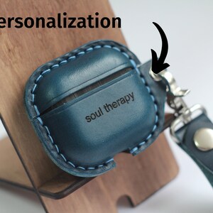 May include: A blue leather Airpods case with a personalized inscription that reads "soul therapy". The case has a blue stitching detail and a silver metal hook.