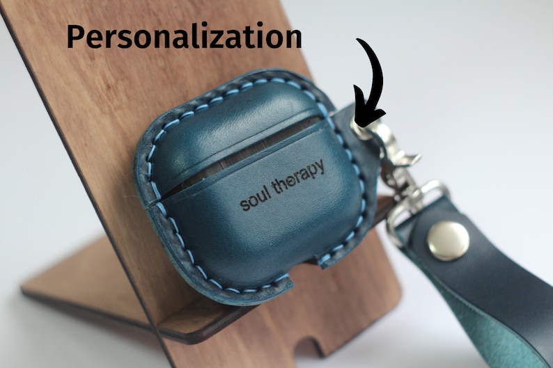 May include: A blue leather Airpods case with a personalized inscription that reads "soul therapy". The case has a blue stitching detail and a silver metal hook.