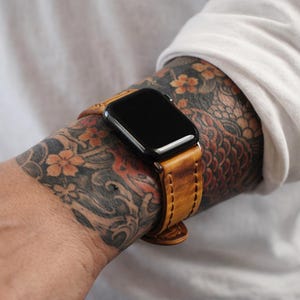 Peut inclure: Une montre intelligente noire avec un bracelet en cuir marron clair est présentée. Le bracelet a un aspect vieilli et usé. La montre est fixée au poignet avec une boucle. Le cadran de la montre est noir et rectangulaire.