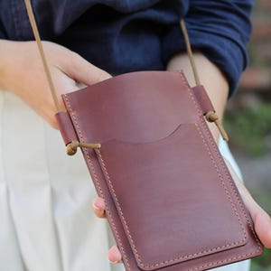 Op de afbeelding: Een bordeauxrode leren crossbodytas met een voorvak en een lange, dunne riem. De tas is rechthoekig met afgeronde hoeken en wordt door een persoon vastgehouden. De tas is gemaakt van glad leer.