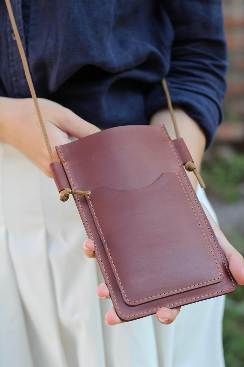 Op de afbeelding: Een bordeauxrode leren crossbodytas met een voorvak en een lange, dunne riem. De tas is rechthoekig met afgeronde hoeken en wordt door een persoon vastgehouden. De tas is gemaakt van glad leer.