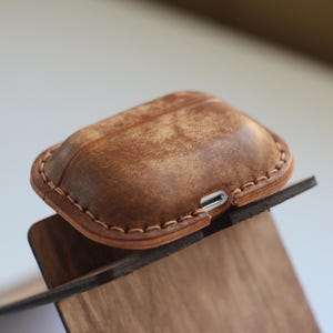 May include: A brown leather case for wireless earbuds with a stitched edge and a charging port.