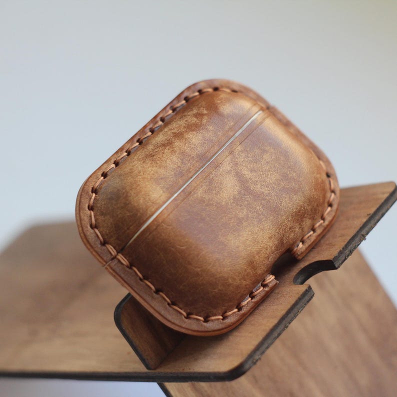 May include: A brown leather case with stitching around the edges. The case is designed to hold a pair of wireless earbuds. The case is sitting on a wooden stand.