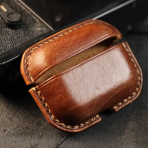 May include: A brown leather case for wireless earbuds. The case is open, revealing the soft brown interior lining.