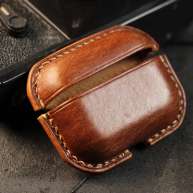 May include: A brown leather case for wireless earbuds. The case is open, revealing the soft brown interior lining.