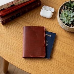 May include: A brown leather passport holder with a blue passport peeking out from underneath. The passport holder is on a wooden table next to a pair of white wireless earbuds and a small potted succulent.