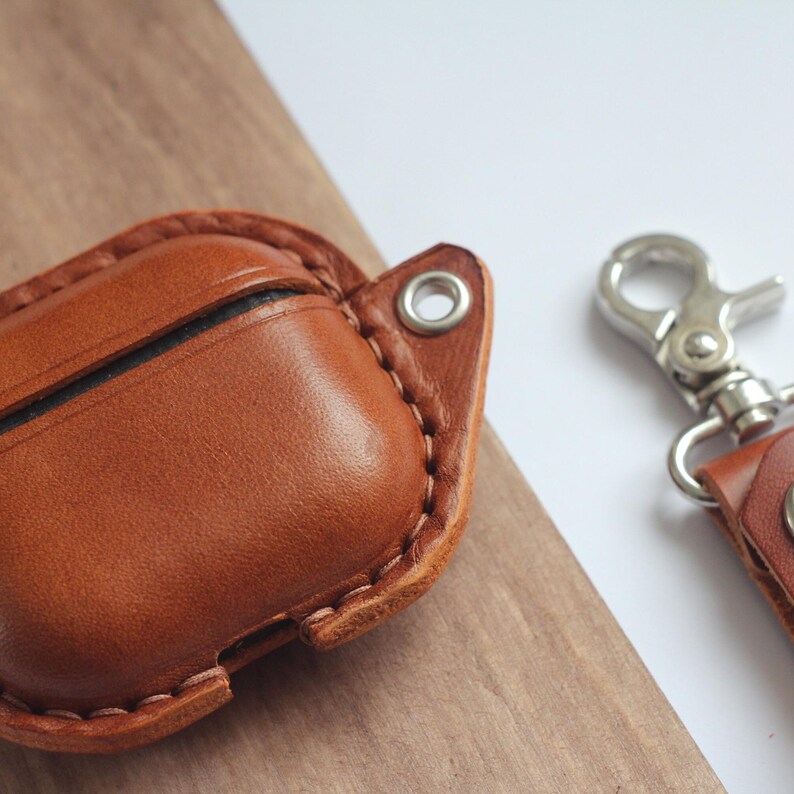 May include: A brown leather case with a stitched edge and a metal loop for attaching a keychain. The case is designed to hold a pair of wireless earbuds.