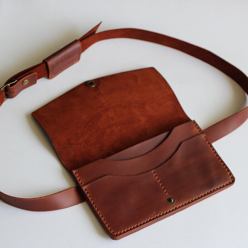 May include: A brown leather belt bag with an adjustable strap. The bag has a flap closure with a press stud and features multiple interior pockets. The leather has a natural grain and a warm, reddish-brown hue. The strap is also brown and has a buckle.