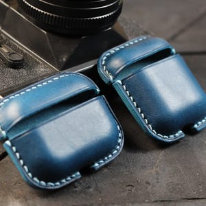 May include: Two blue leather cases with white stitching for wireless earbuds. The cases are made of a soft, supple leather and have a classic, elegant design.